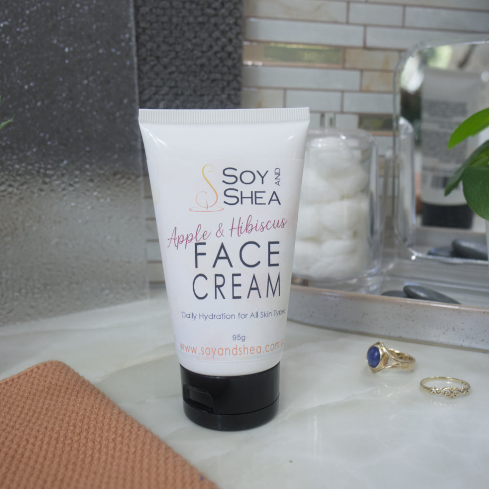 A white squeeze tube of Soy and Shea Face Cream with a black cap, placed on a marble countertop with a peach towel, rings, and a glass jar of cotton pads in a bathroom setting.