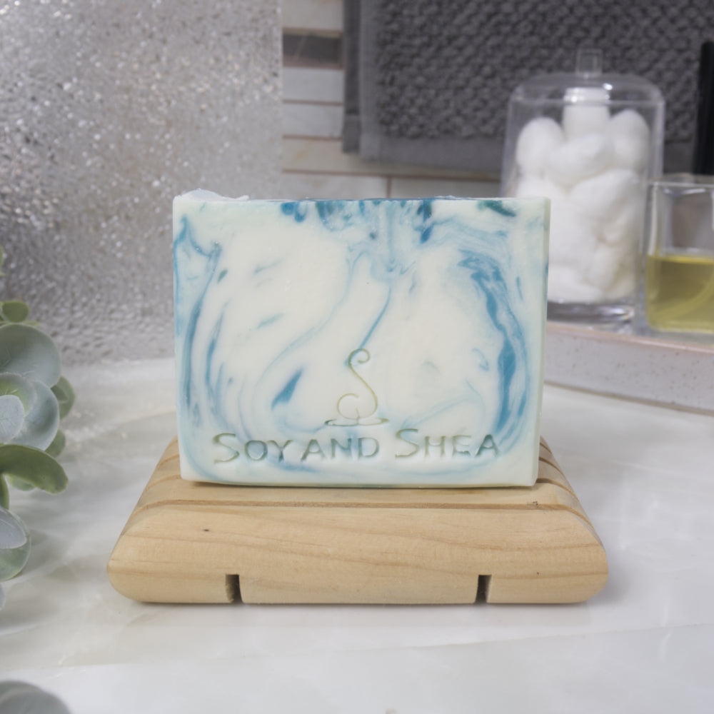 A soap bar sits on a wooden soap rack on a bathroom counter top with a blurred background. The soap has a white base with teak swirls of colour throughout.