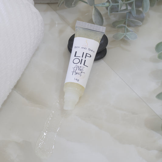 A tube of Lip Oil lies on a marble tile surface next to a white towel and some eucalyptus leaves. The tube is clear with a white product label and filled with an off while Lip oil. The cap is removed and a smear of Lip oil is on the bench showing the translucent effect it has