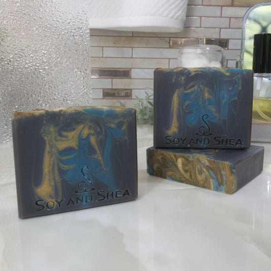 Three bars of Soap and Shea soap on a bathroom counter with a tiled wall in the background. One soap lays flat on the surface showing a top that had dark gold and blue swirls. A second soap stands upright on this soap with a third to the front-left. These soaps are dark blue with bright blue and gold swirls of colour dropped through.