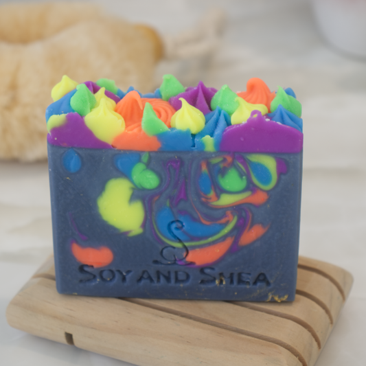 A dark blue soap bar wiht neon coloured drops and piped top sits on a wood soap dish with a blurred background