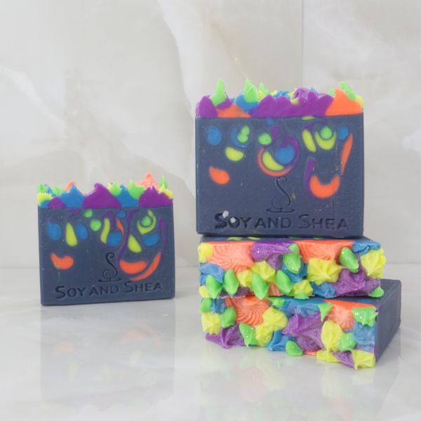 A collection of Aurora Bloom Soap Bars are displayed on a marble tile background. Two soaps lay flat on the tile stacked up each other showing the tops of the soap with neon coloured piping. A third soap stands up right on these showing a dark blue soap with neon colour drops throughout. A fourth soap is in the background with the same colours but different pattern