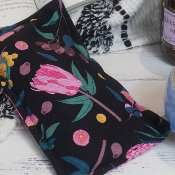 A small rectangular jeat pack lies on a wooden bench. It is a close up shot showing the fabric pattern of a black background with yellow wattle, pink gum flowers and gum nuts.
