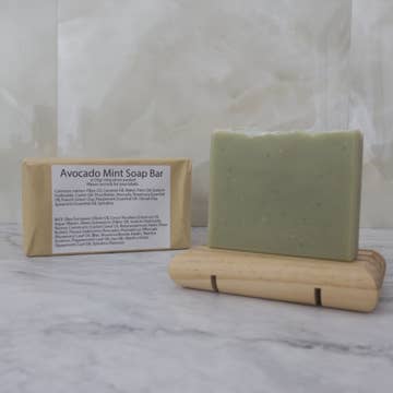 Pale green soap bar on a wooden stand with a brown paper package labeled 'Avocado Mint Soap Bar' on a marble background