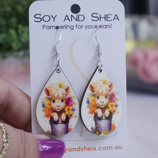 A hand holds a white card branded with SOY AND SHEA with a pair of earrings hanging on hooks. The earrings are teardrop shaped featuring an illustration of a baby highland cow sitting in a grey bucket surrounded by flowers on white background