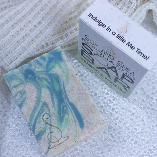A cream rectangular bar of soap with pale and dark green swirls and brown speckles is laying in water. Beside it is a cream face wash and a box that the soap comes in. The box has a pale green border around its base and is white at the top.