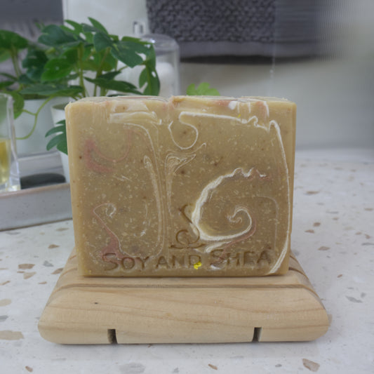 A bar of soap stands on a wooden soap rack next to a plant on bathroom counter top. the soap has a golden base colour with swirls of white and bronze