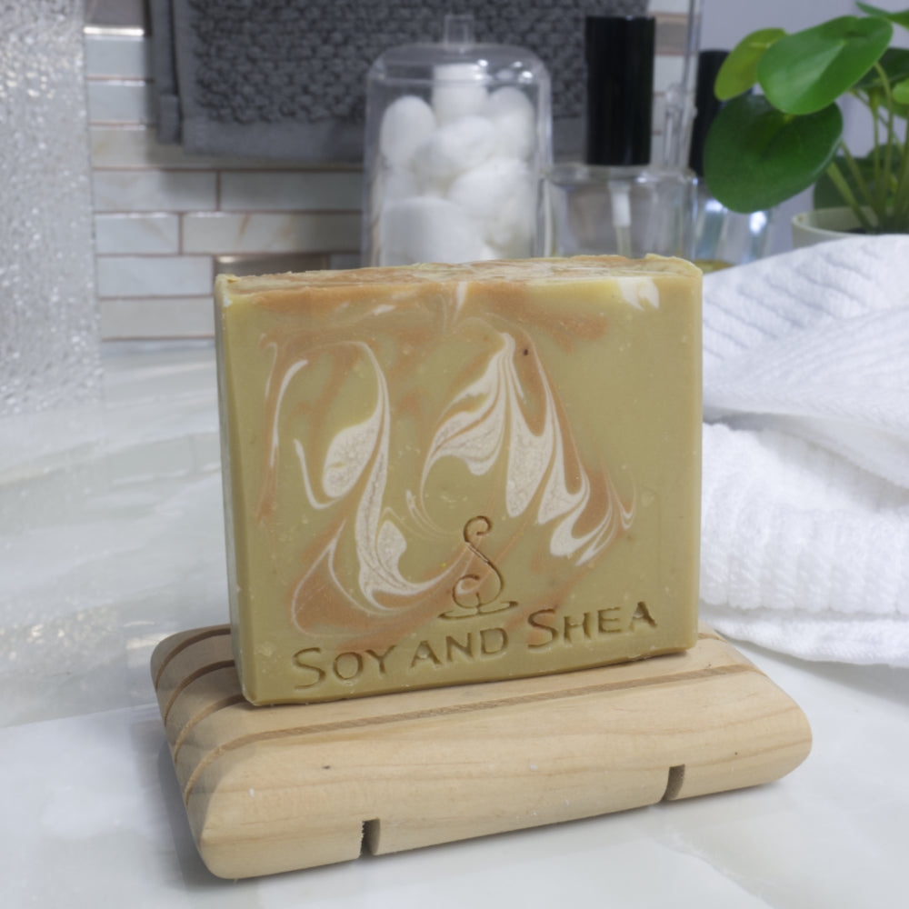 A bar of soap stands on a wooden soap rack next to a white towel on bathroom counter top. The soap has a golden base colour with swirls of white and gold