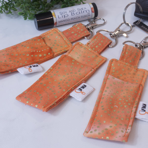 A selection of three clip balms showing variations in fabric design, lie on a wooden side with the middle Clip Balm attached to a car key. A lip balm lays near-by along with some greenery. The Clip balms are a vibrant orange with gold dots scattered over them