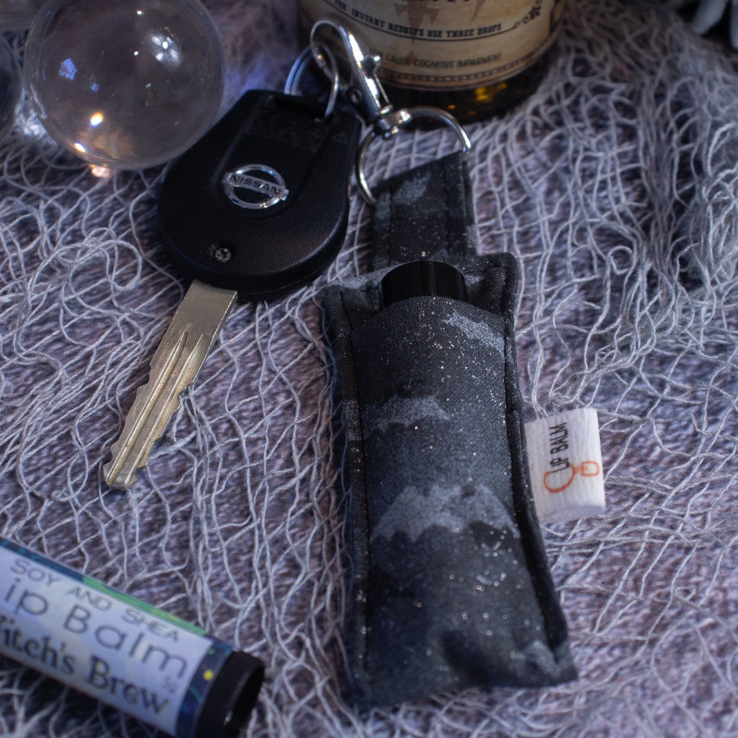 A Lip Balm holder lies on a grey mesh fabric attached to a car key with a lobster claw clasp. Next to it is a lip balm and glass orb. The Clip Balm is a dark grey with images of solid light grey and black bats randomly scattered over the fabric