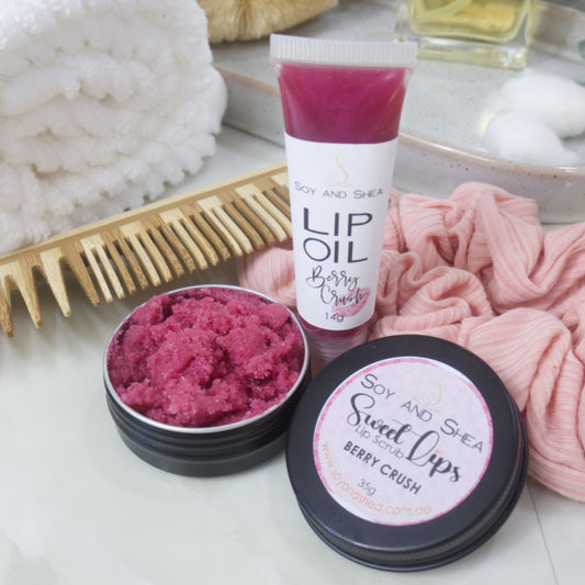 A small black tin with the lid removed revealing a cerise Lip Scrub sits on a marble tile surface next to a small, clear tube of cerise coloured Lip Oil. This rests againsts a wooden comb and pink scrunchie.