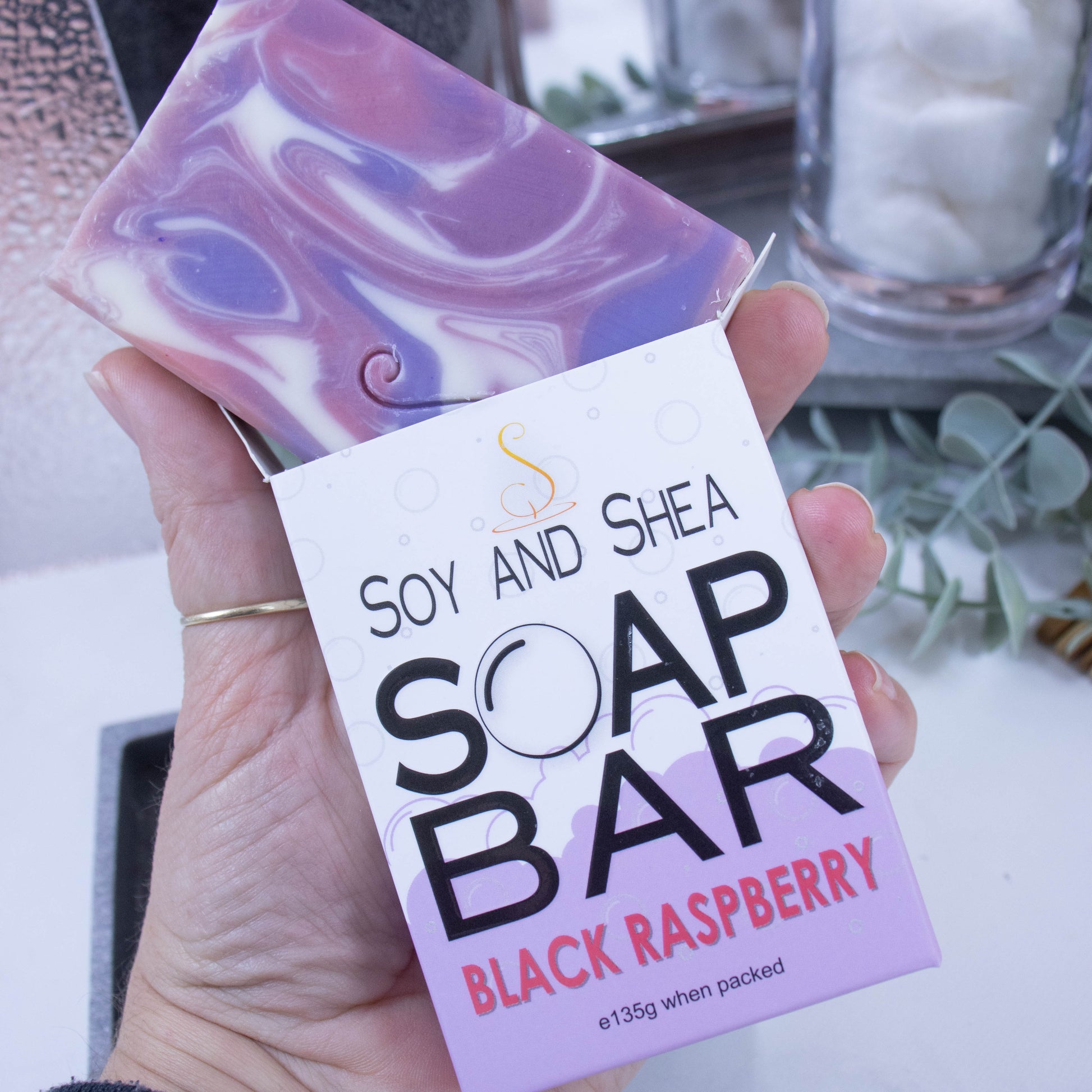 A hand is holding onto a box that has a bar of soap coming out the top.  The lower half of the box has a mauve bubble shaped border and the top is white with grey bubbles.  The box shows it is soap and the scent.  The soap is rectangular in shape and features a swirling pattern of white, pink  and purple.