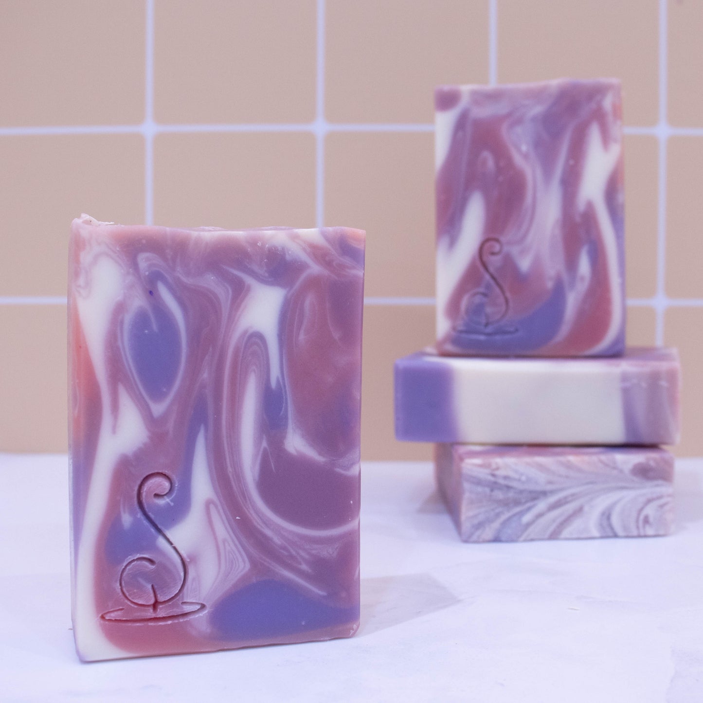 Four rectangular bars of soap sit on a marble bench each showing various design patterns from the same batch of soap. All soaps feature a swirling pattern of white, pink and purple soap but all have slight differences