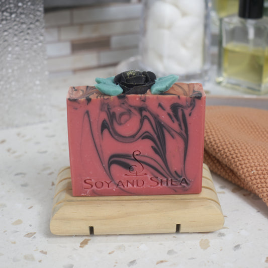 A red bar of soap with black swirls sits on a wooden soap dish on a bathroom counter with a blurred background. The soap top features a black rose piped with soap and surrounded by green leaves