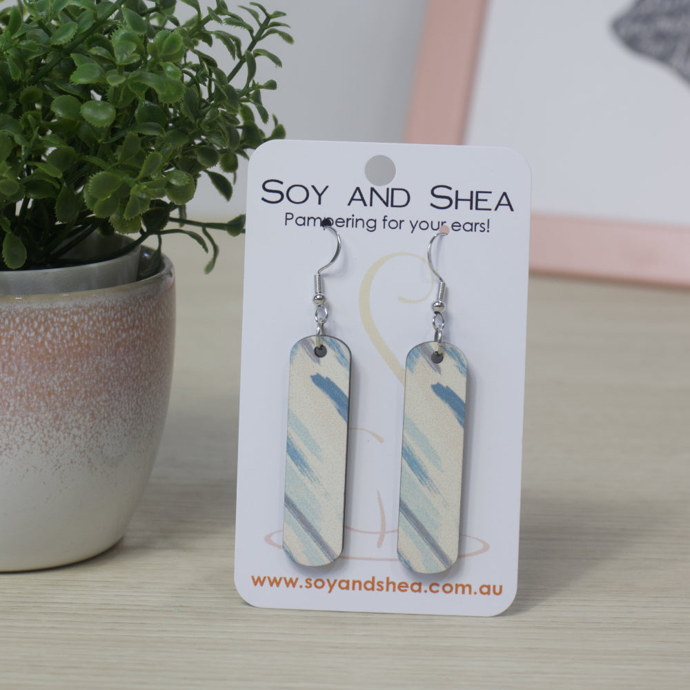 Long rectangular earrrings with rounded corners hang on a branded Soy and Shea card next to plant. The earrings feature a design of brush strokes in various blue shades