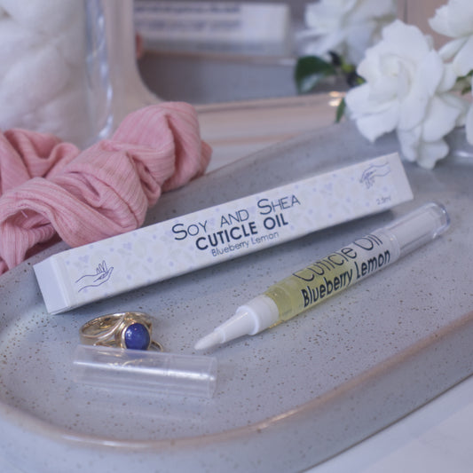 A clear, narrow tube with the lid removed showing a brush tip lies on a stone tray beside a cardboard box labelled CUTICLE OIL and a set of gold rings. In the background is a bathroom counter top with a pink scrunchie, small mirror, and a some white flowers.
