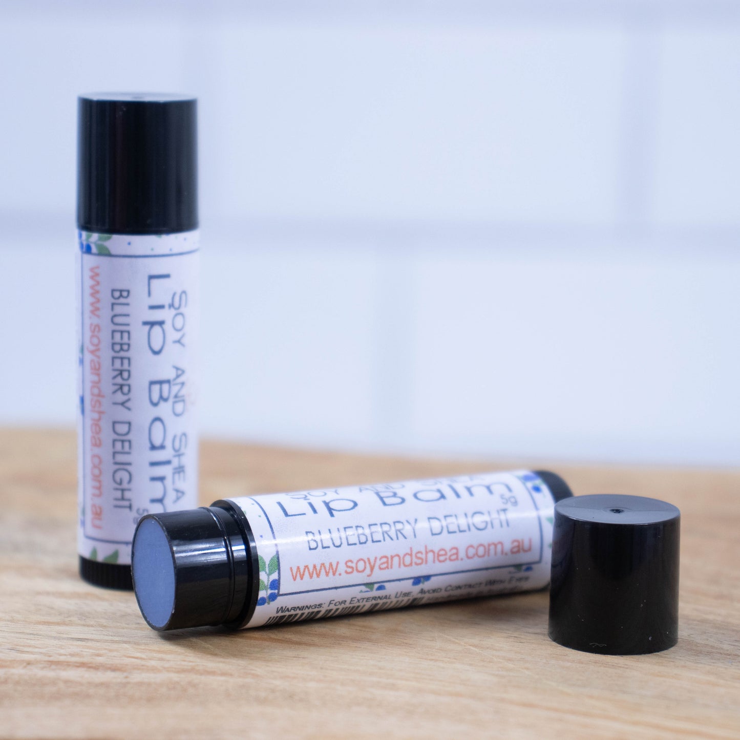 Two black tubes of lip balm are displayed on a wooden surface with blurred tiled background. One tube stands on its end while the other lays flat in front with the cap removed revealing a blue balm. The label is white with a random pattern of blueberries with green leaves.