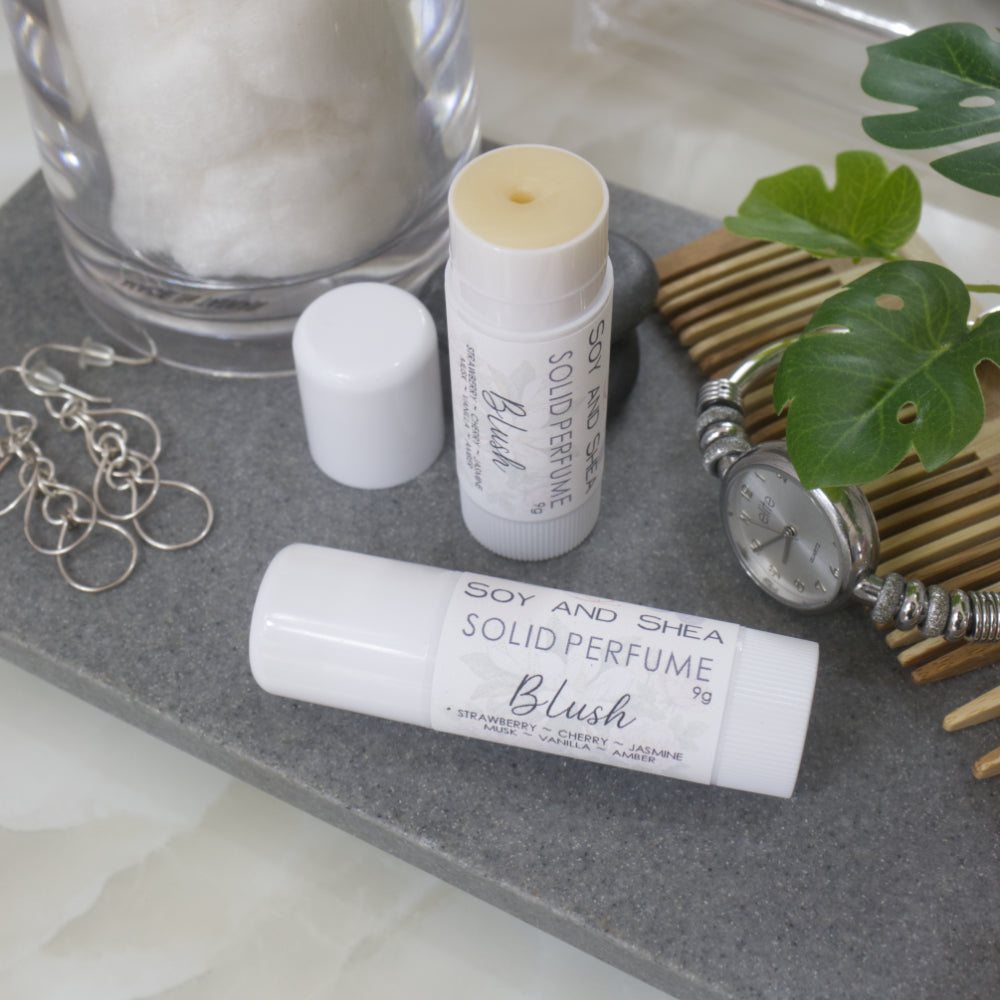 Two white tubes of Solid Perfume are displayed on a grey stone tray along with silver earrings, watch and greenery. One tube lays flat displaying the fragrance notes while another tube stands upright behind it with the lid removed and to the left revealing a beige balm