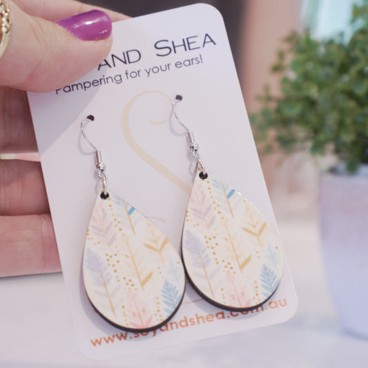 A hand holds a white card branded with SOY AND SHEA with a pair of earrings hanging on hooks. The earrings are teardrop shaped with boho style feathers in pastel shades of blue, purple and pink with gold highlights