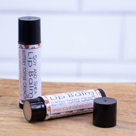 Two black tubes of lip balm are displayed on a wooden surface with blurred tiled background. One tube stands on its end while the other lays flat in front with the cap removed revealing a light brown balm. The label is white with a random pattern of caramel toffees.