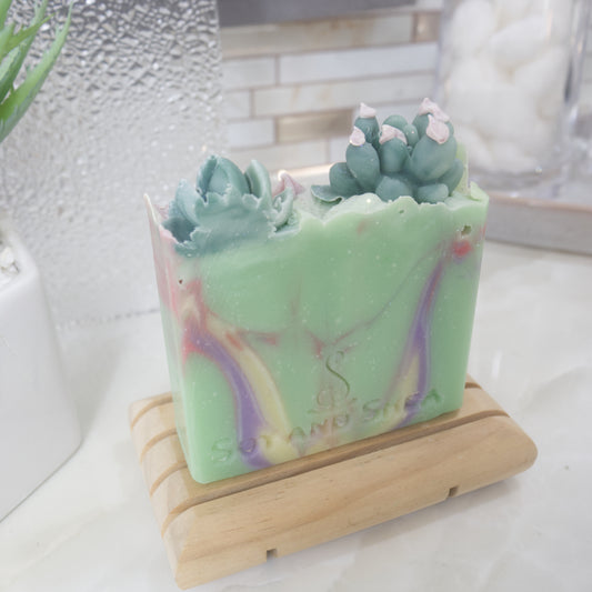 A close up shot of soap bar standing on a wooden soap rack with a blurred background. The soap is bright green with swirls of purple, yellow and red. On the top of the soap is a selection of Cacti embellishments.