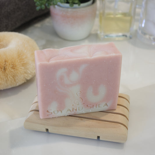 An overhead shot showing a pale pink soap bar with white swirls sitting on a wooden soap dish on a bathroom counter top. In the background a body brush and bottle of perfume can be seen.