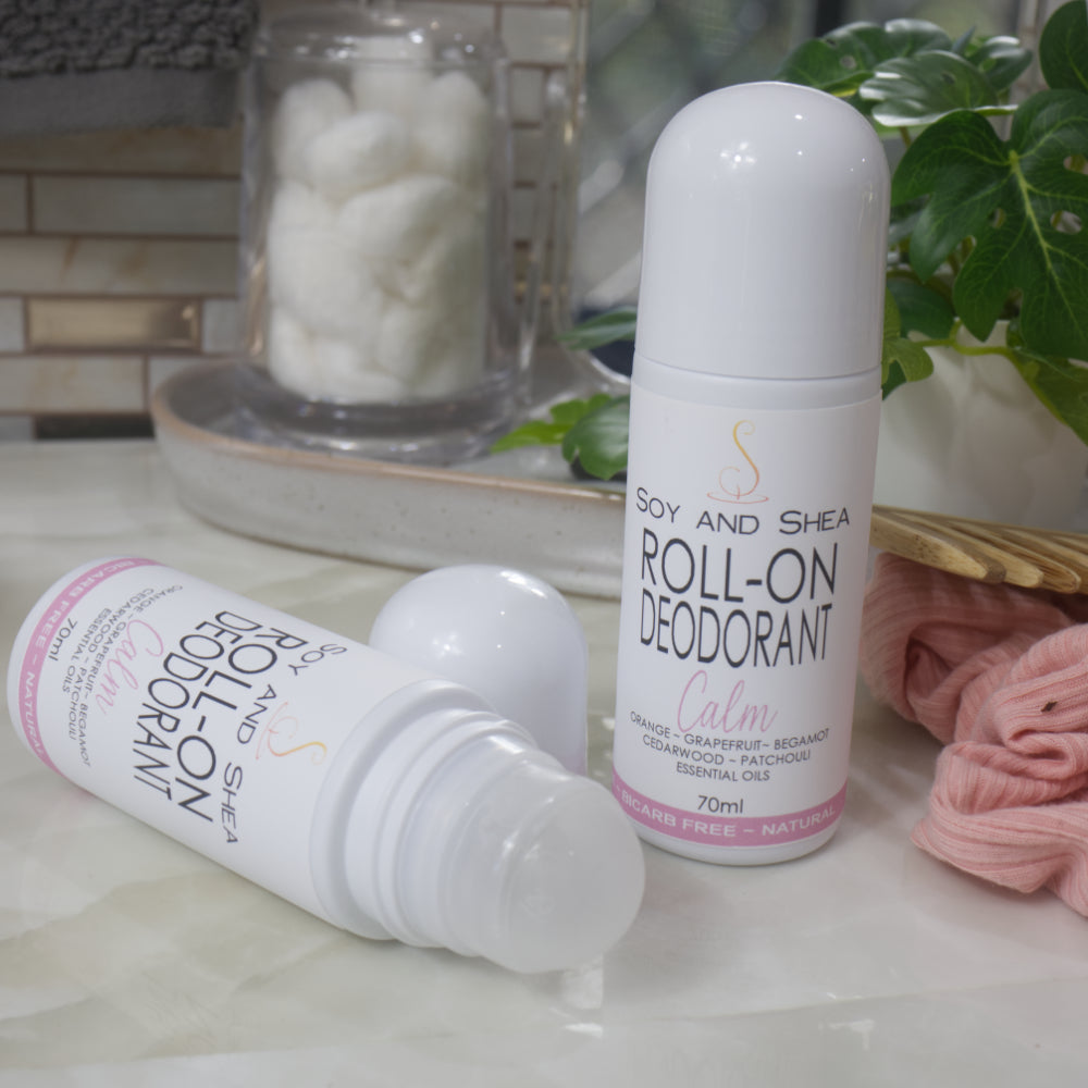 Two bottles of Roll-On Deodorant are displayed on a bathroom counter top with a jar of cotton balls, hair tie and greenery. One bottle stands upright with a domed cap, while another lays flay with the cap removed showing a ball roller in the top