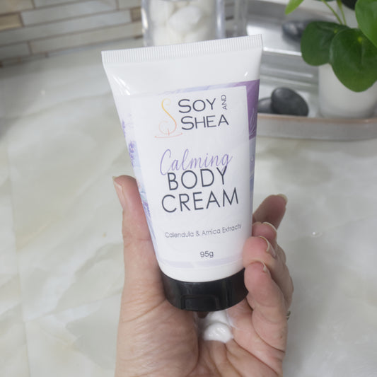 A hand holds a white tube with black cap labelled as Calming Body Cream with calendula and Arnica Eztracts. In the palm of the hand is a dollop of white cream that has been squeezed out of tube.