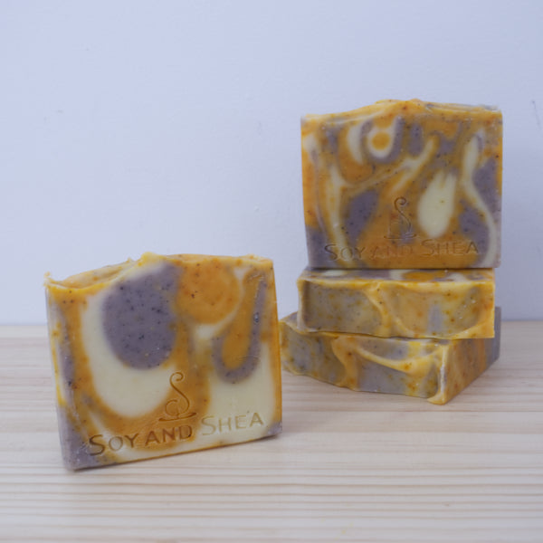 A selection of Calming Citrus Soap bars rest on a wooden surface with white background. Two soaps lie flat, stacked upon each other showing a textured top. A third soap with purple, yellow and white swirls stands on top of the soap stack with a fourth soap in front of these soaps. This soap shows a variation in pattern.