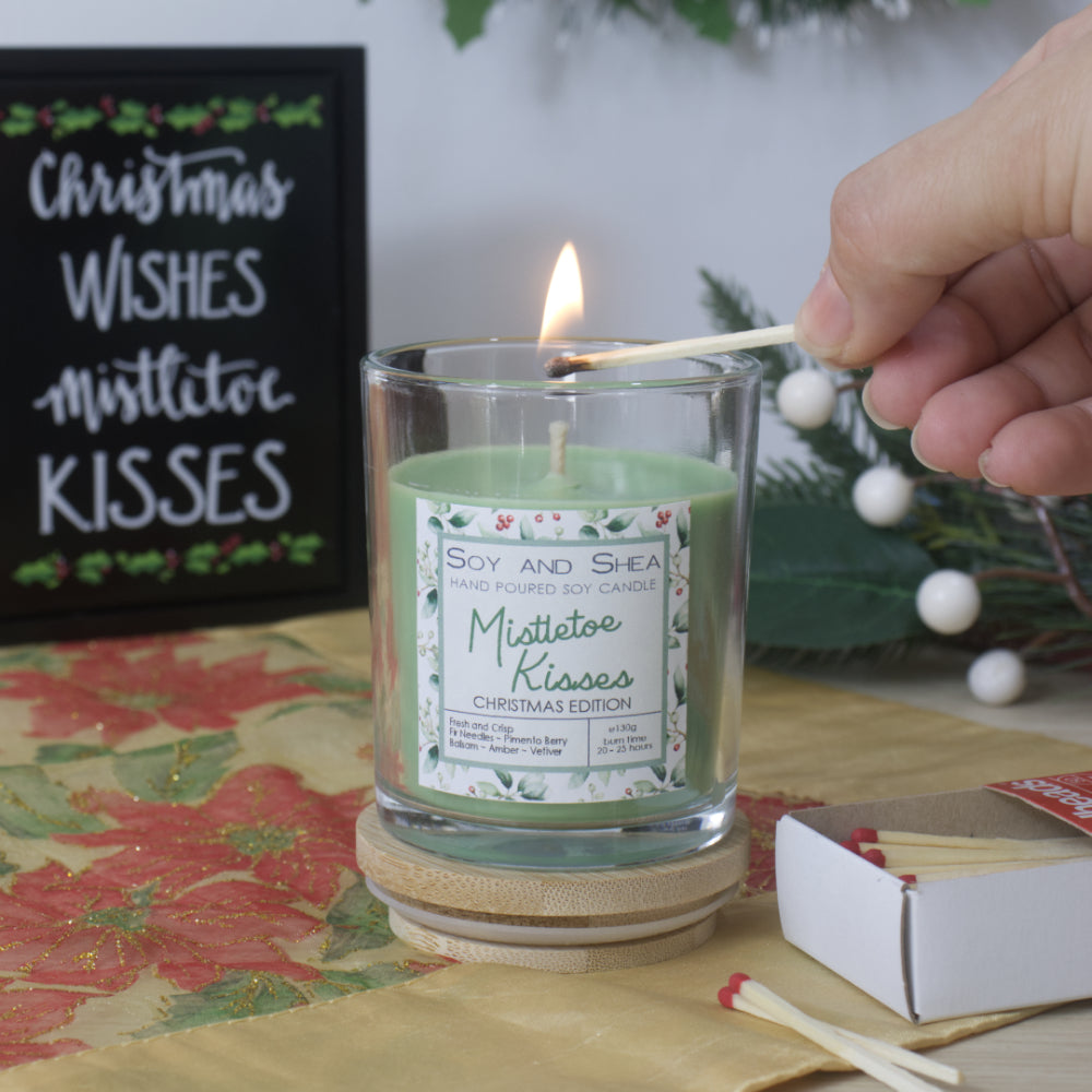 A hand reaches in to light a candle in a clear glass filled with green wax. The jar stands on a wooden lid and is surrounded by a box of matches with Christmas greenery