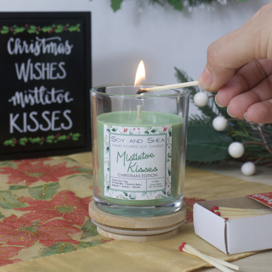 A hand reaches in to light a candle in a clear glass filled with green wax. The jar stands on a wooden lid and is surrounded by a box of matches with Christmas greenery