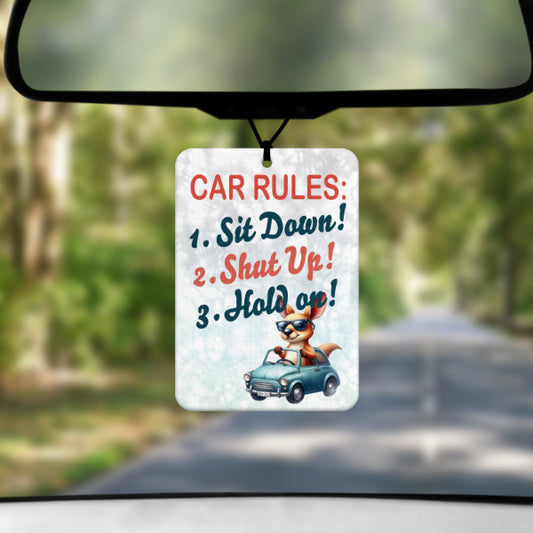 A mock up image showing a view through a car windscreen with a long tree lined road. From the rearview mirror hangs an air freshener which list CAR RULES. SIT DOWN. SHUT UP. HOLD ON. In the lower corner is a cartoon image of a large kangaroo driving a small blue car with the roof down
