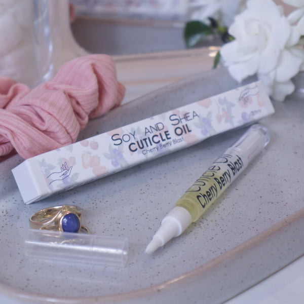 A clear, narrow tube with the lid removed showing a brush tip lies on a stone tray beside a cardboard box labelled CUTICLE OIL and a set of gold rings. In the background is a bathroom counter top with a pink scrunchie, small mirror, jar of cotton balls and a some white flowers.
