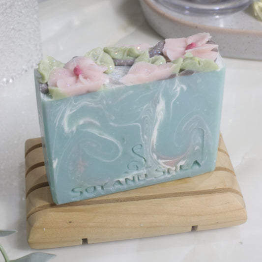 A bar of soap stands on a wooden soap dish on a bathroom counter top. The soap has a pale blue base colour with swirls of white and pink. The top has pink flowers and green leaves piped with soap.