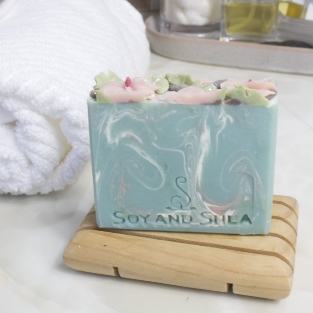 A bar of soap stands on a wooden soap dish on a bathroom counter top next to a white rolled-up towel. The soap has a pale blue base colour with swirls of white and pink. The top has pink flowers and green leaves piped with soap