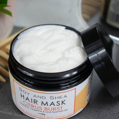 An overhead shot of a black jar with the lid resting on its side revealing a white cream inside, sits on a dark grey tile surface against a blurred background of a bathroom counter top. The jar features a white label with yellow abstract design with text HAIR MASK CITRUS BURST.