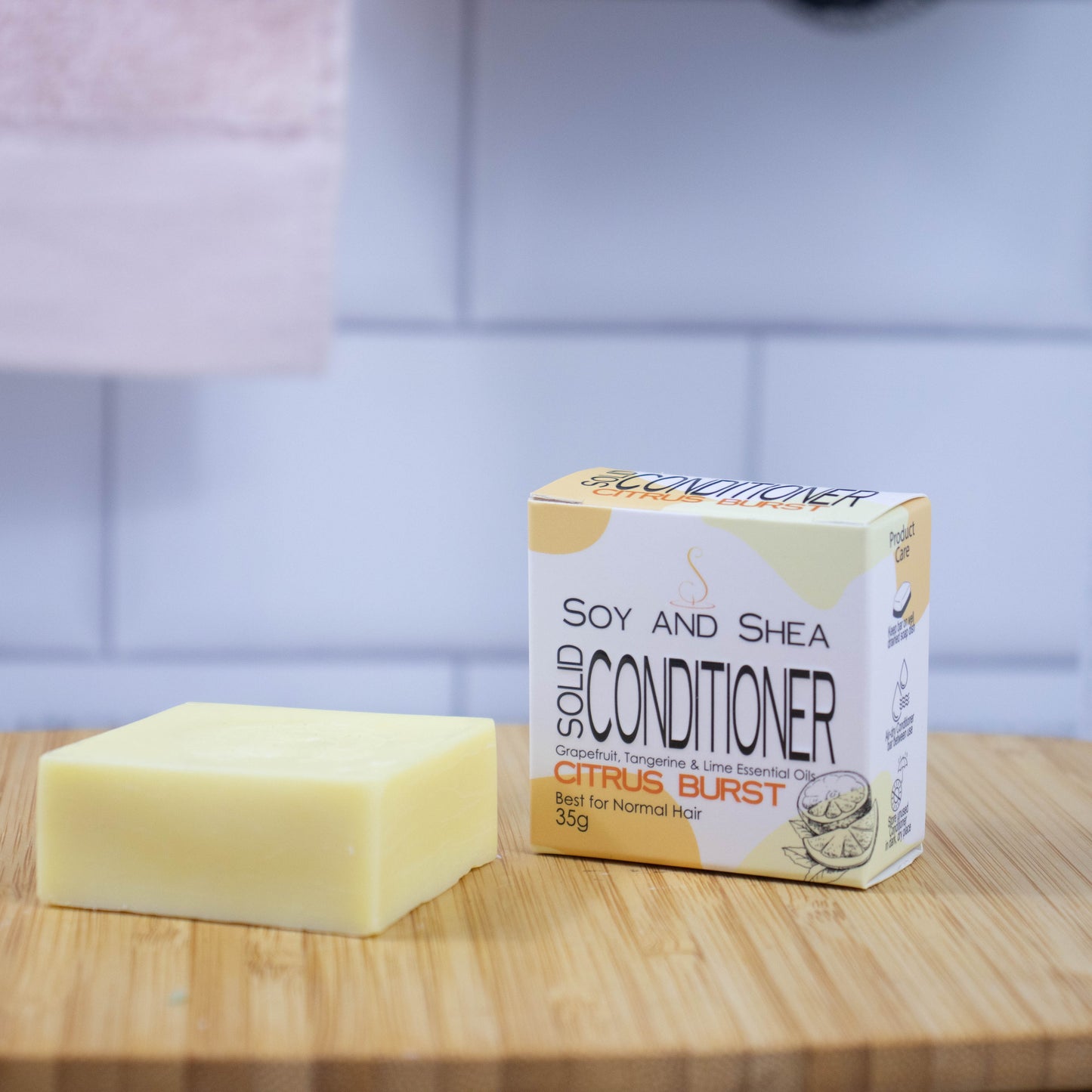 On a wooden tray sits a yellow bar of conditioner next to a white box with a yelllow abstract pattern reading SOLID CONDITIONER against a blurred tiled background.