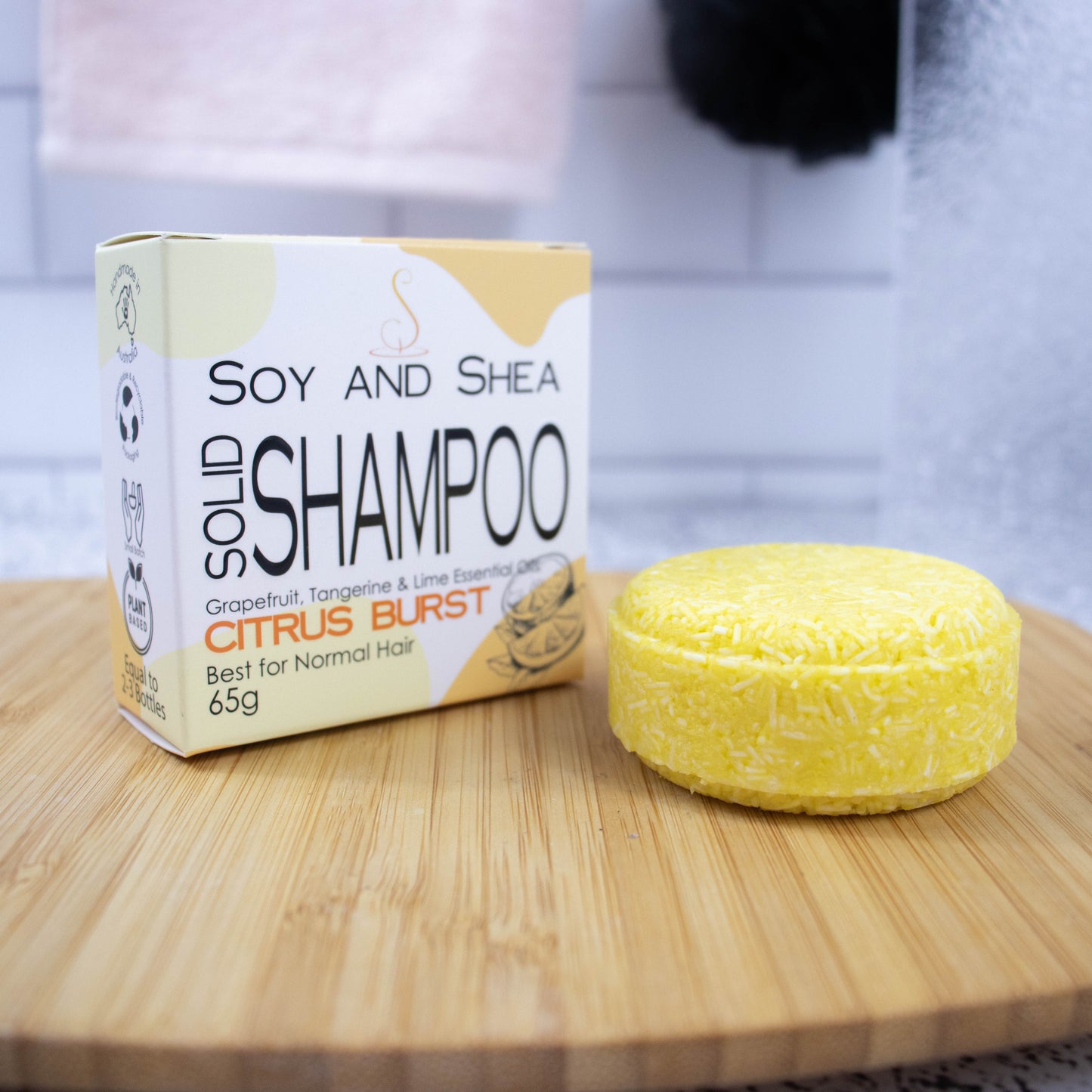 A white box with yellow abstract pattern and the text SOLID SHAMPOO CITRUS BURST stands upright on a wooden tray with a blurred bathroom background. Next to the box sits a round bar of solid shampoo. The bar is bright yellow with small white lines throughout.