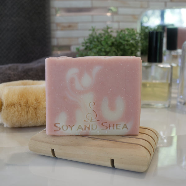 A pale pink soap bar with white swirls sits on a wooden soap dish surrounded by a grey towel, plant and bottle of perfume in front of a mirror.