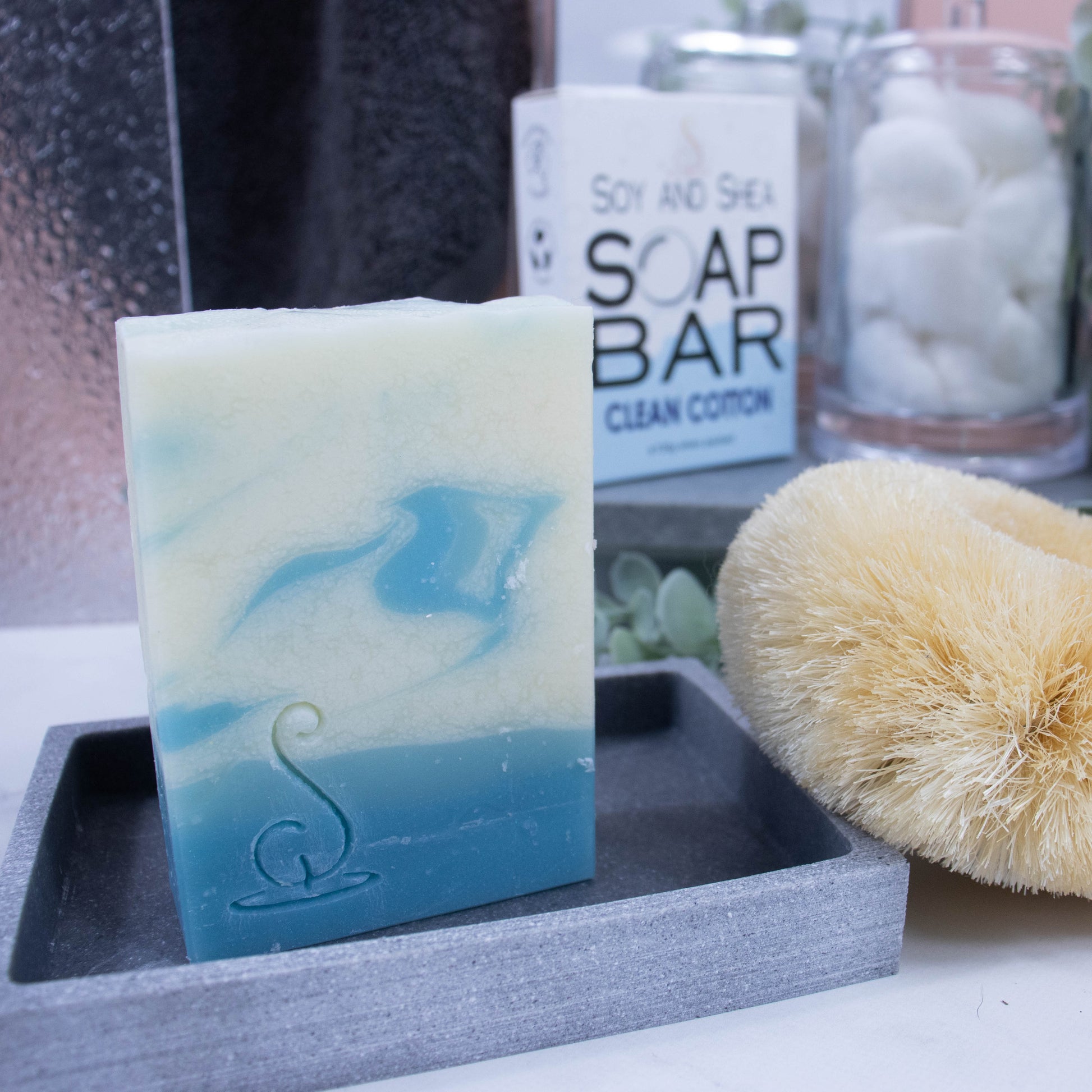 A close up look of the soap which has layers of colour including two shades of pale blue at the base and the top layer being white with whisps of the blues swirled through. The soap is rectangular in shape and is taller than wide. It is sitting on a stone soap dish and behind it is the box the soap came in. The lower portion of the box has a bubble border coloured in pale blue whereas the top is white with small grey bubbles. The box advises it is soap and the scent.