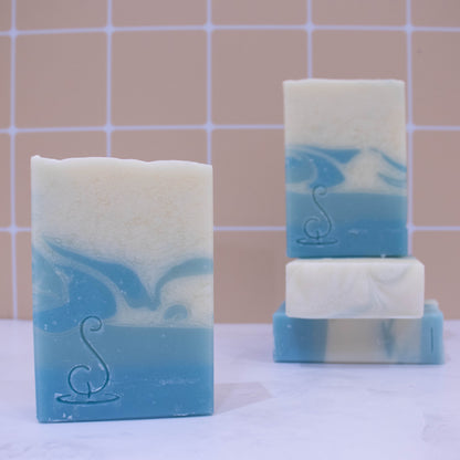 Four rectangular bars of soap sit on a marble bench each showing various design patterns from the same batch of soap. All soaps feature a base of two layers of pale blue soap, one darker than the other and the top layer of the soap is white with whisps of the blue swirled through.  All soaps have a similar pattern but all have slight differences