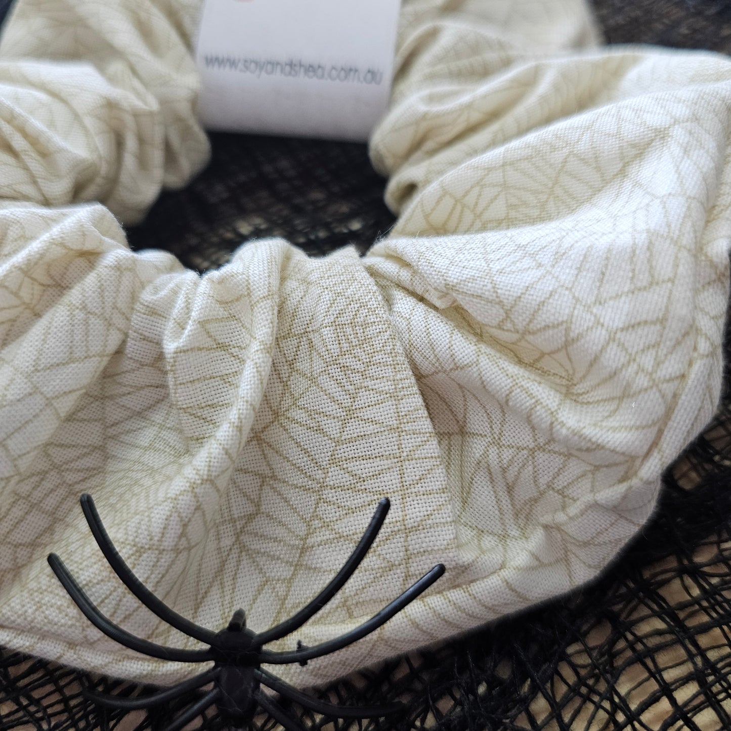 A detailed view of the Cobwebs Halloween Scrunchie. It shows a cream coloured scrunchie with a gold cobweb design all over
