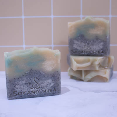 A collection of soaps sit on a white marble surface against a pale peach tile background. In the background, two soaps lay flat but stacked upon each other revealing a textured cream top. A third soap stands on stop of this stack with a fourth soap in the foreground. The Soaps have two layers with the base being grey with black flecks and the top is cream with green swirls.