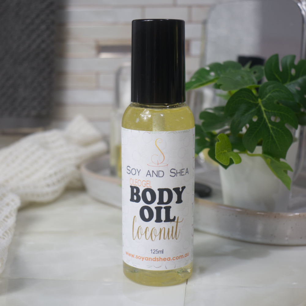 A clear bottle with black lid stands on a bathroom counter with a plant in the background. The bottle is filled with a clear, golden gel and features a product label identifying it as Body Oil in Coconut Scent