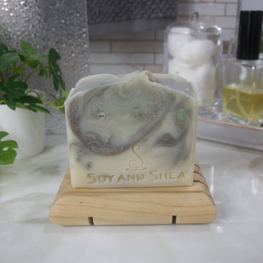 A bar of soap stands on a wooden soap dish on a bathroom counter top along with a plant, a jar of cotton balls and bottle of perfume. The soap has a cream base colour with brown and light green swirls throughout.