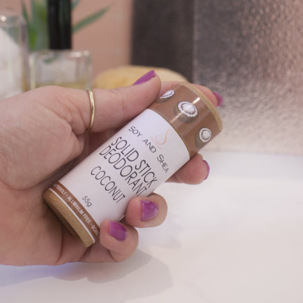 A hand holds a cardboard tube labelled as SOLID STICK DEODORANT against a blurred bathroom background.