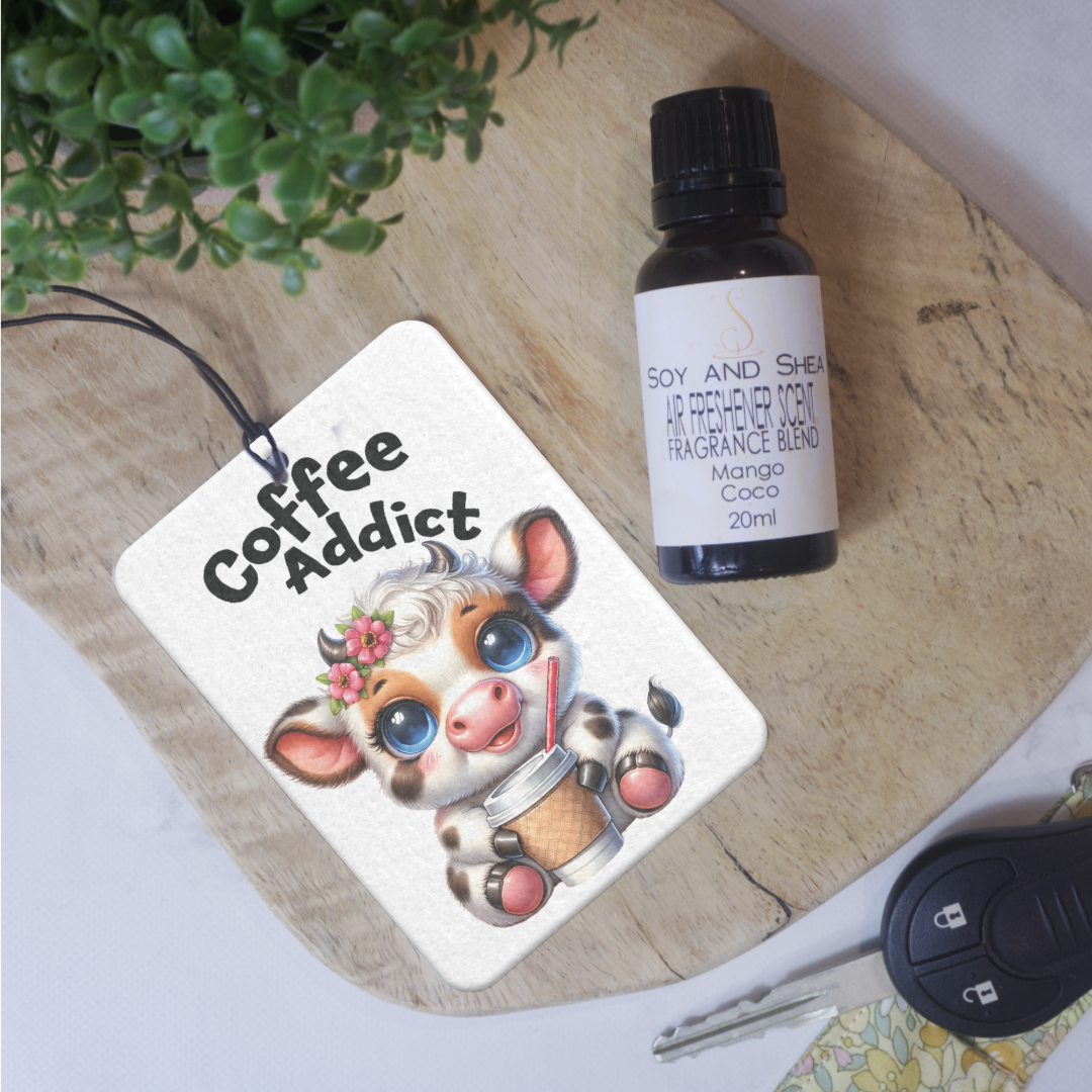 A felt air freshener lies on a wooden surface with an image of a baby cow holding a coffee cup with straw and the words COFFEE ADDICT. Next to it is a bottle of scent and a car key
