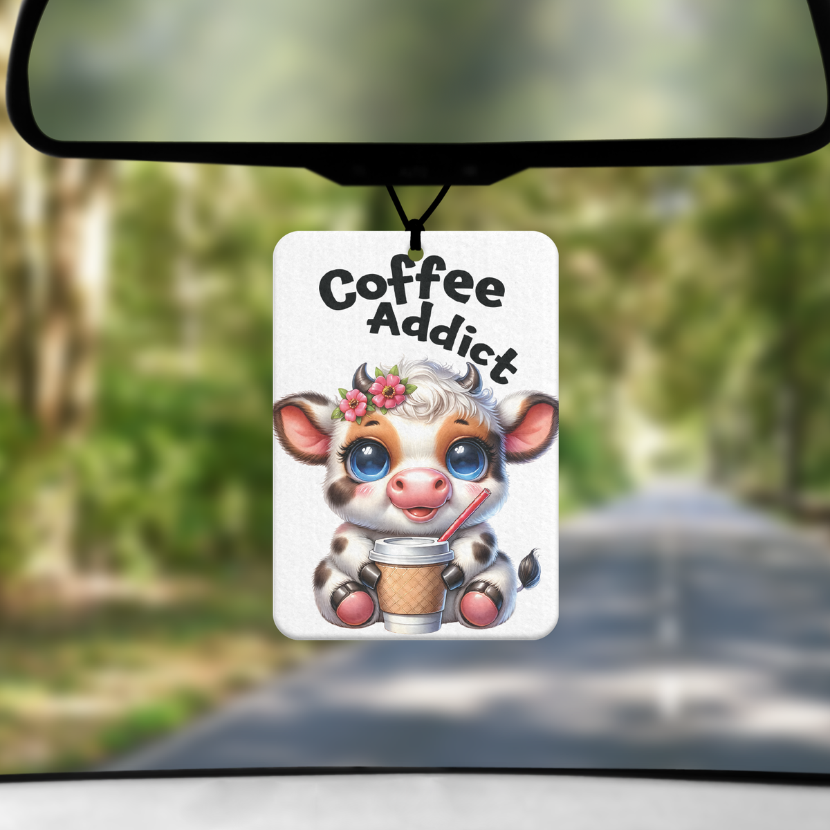 A mock up image showing a view through a car windscreen with a long tree lined road. From the rearview mirror hangs an air freshener with a cartoon image of black and white cow with flowers in hair drinking a cup of coffee with the words COFFEE ADDICT