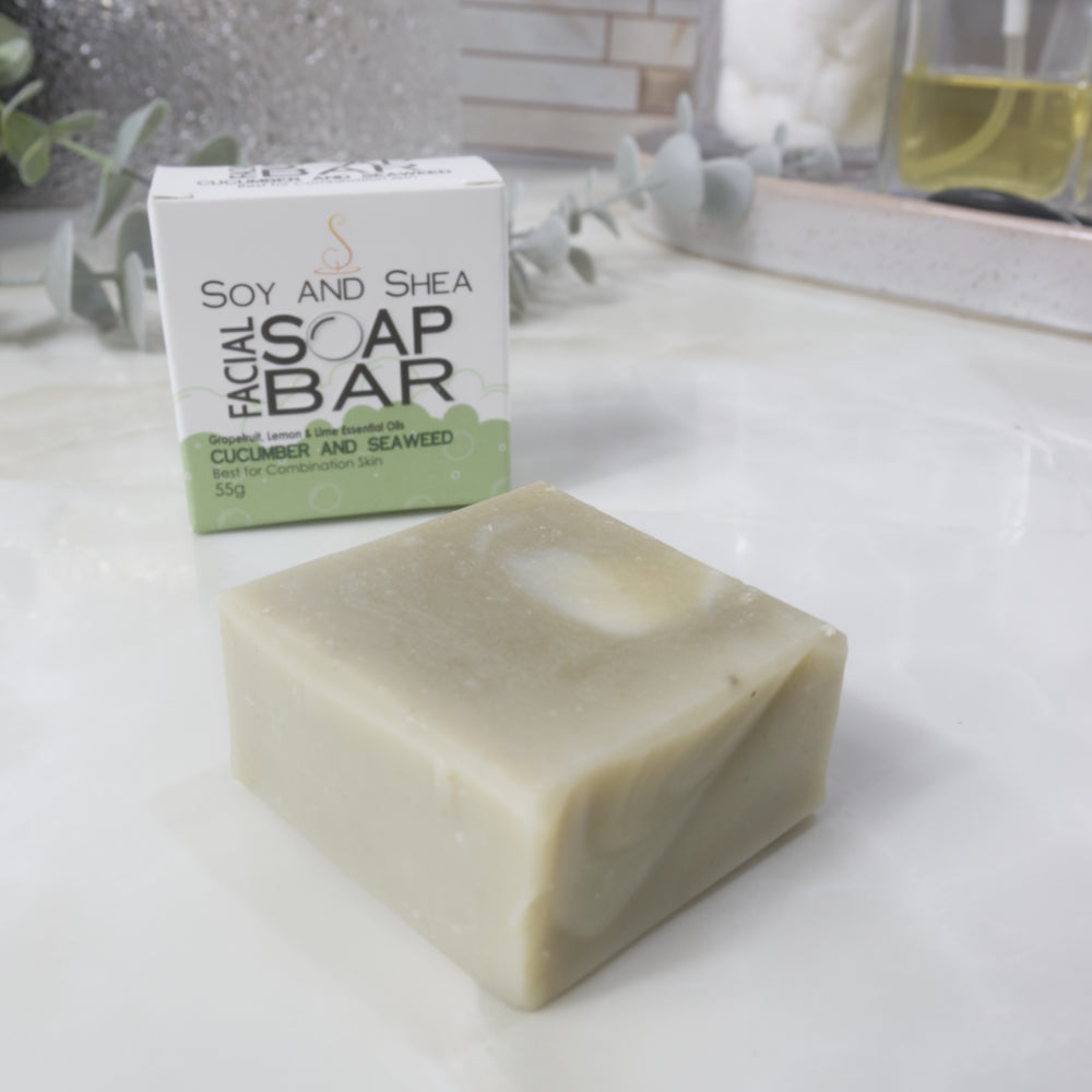 A small square bar of pale green soap in the foreground with its white and light green box behind it on a light countertop. The box reads 'Soy and Shea Facial Soap Bar' and 'Cucumber and Seaweed'.