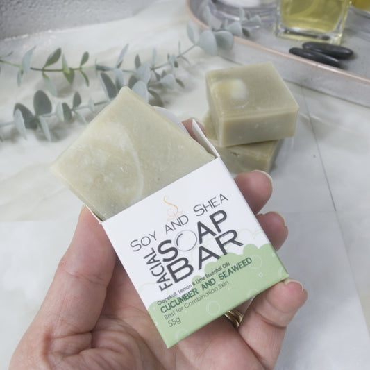 A hand holds a white and green box labelled as Facial Soap Bar with Cucumber and Seaweed. The top of the box is open and a bar of pale green soap is shown coming out the top. On the counter top is a stack of two more soaps along with some greenery